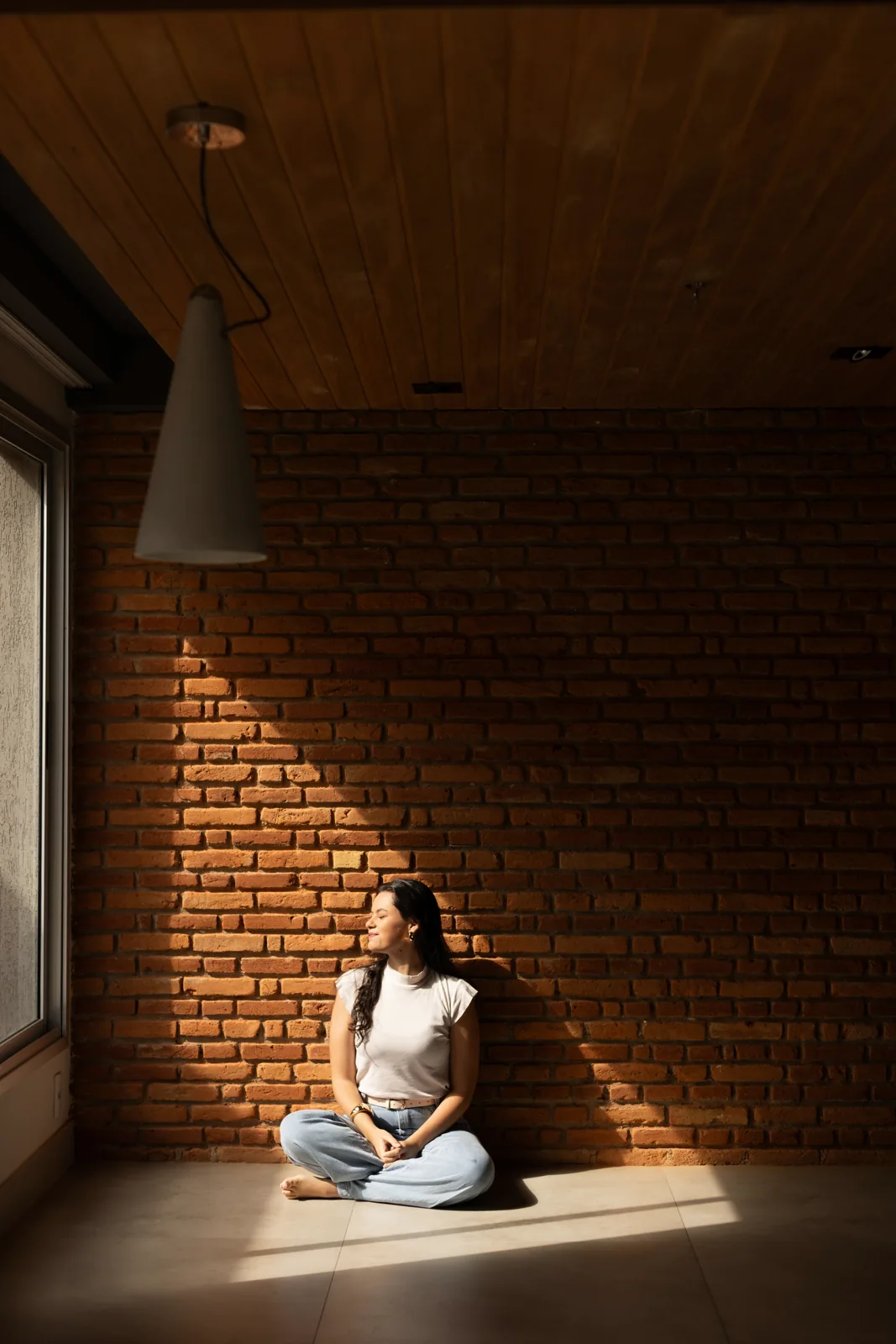 Raquel sentada no chão, junto a uma parede de tijolos iluminada pelo sol