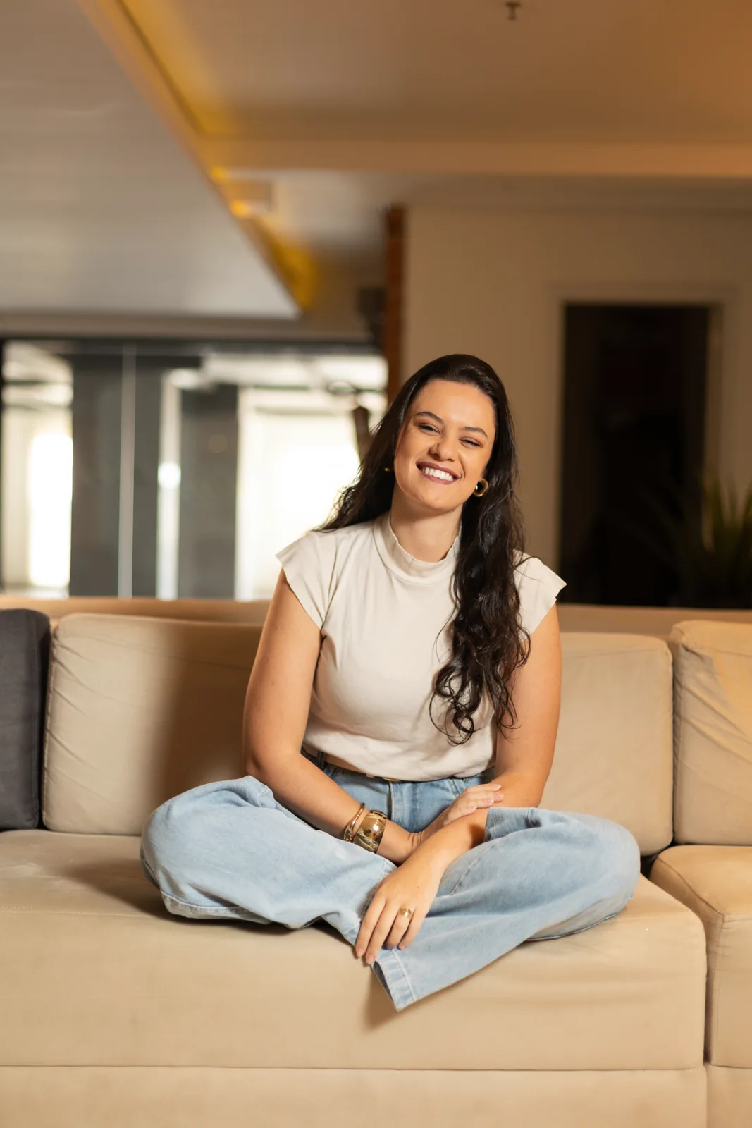 Raquel Lopes sorrindo, sentada no sofá em ambiente acolhedor